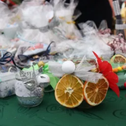Ręcznie zdobione świeczki w słoiczkach i upominki zapakowane w celofan, ozdobione suszonymi pomarańczami, wstążkami i sztucznymi kwiatami, prezentowane na zielonym stole w Gliwicach.