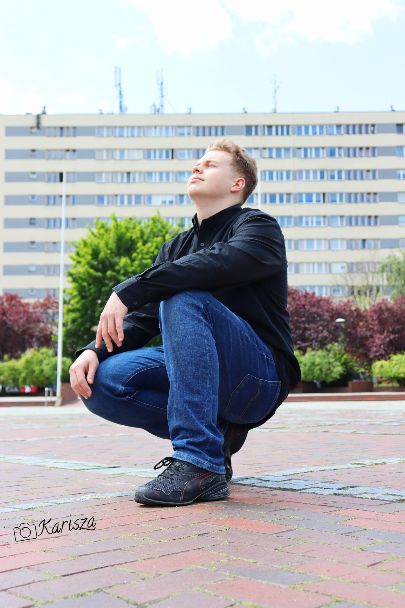 Młody mężczyzna w czarnej koszuli i jeansach kuca na kostce brukowej, spoglądając w górę na tle budynku z licznymi oknami i zielenią.