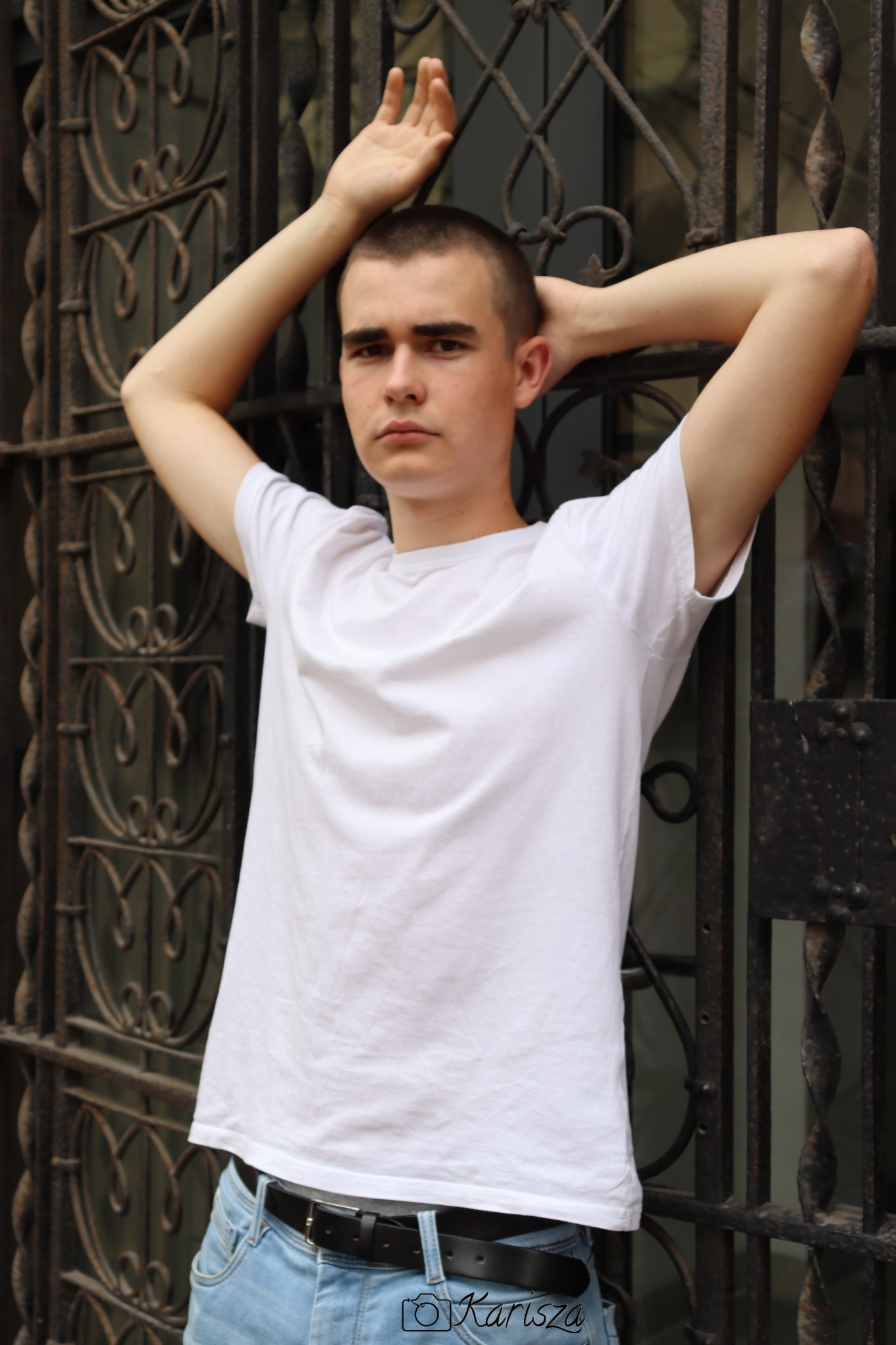 Młody mężczyzna w białym t-shircie i jeansach, oparty o ozdobną, metalową bramę, z rękami uniesionymi nad głową i skierowanymi do góry.