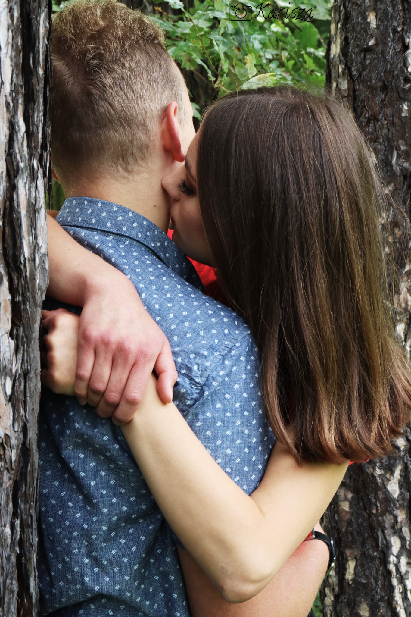 Para przytulona do drzewa, kobieta całuje mężczyznę w szyję, widoczne fragmenty drzew, ubrania i włosów.