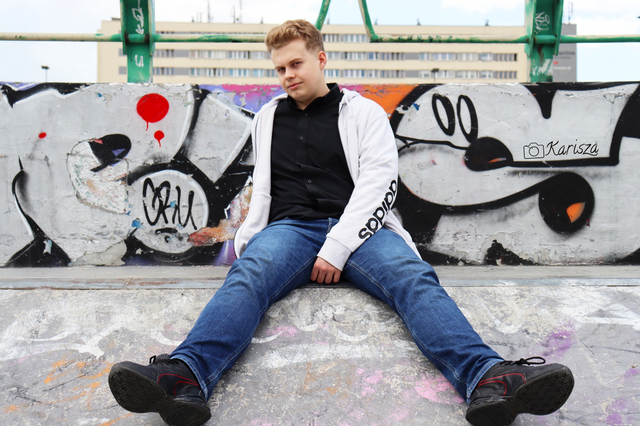 Młody mężczyzna w sportowej bluzie i jeansach siedzi na betonowej ścianie skateparku z graffiti.