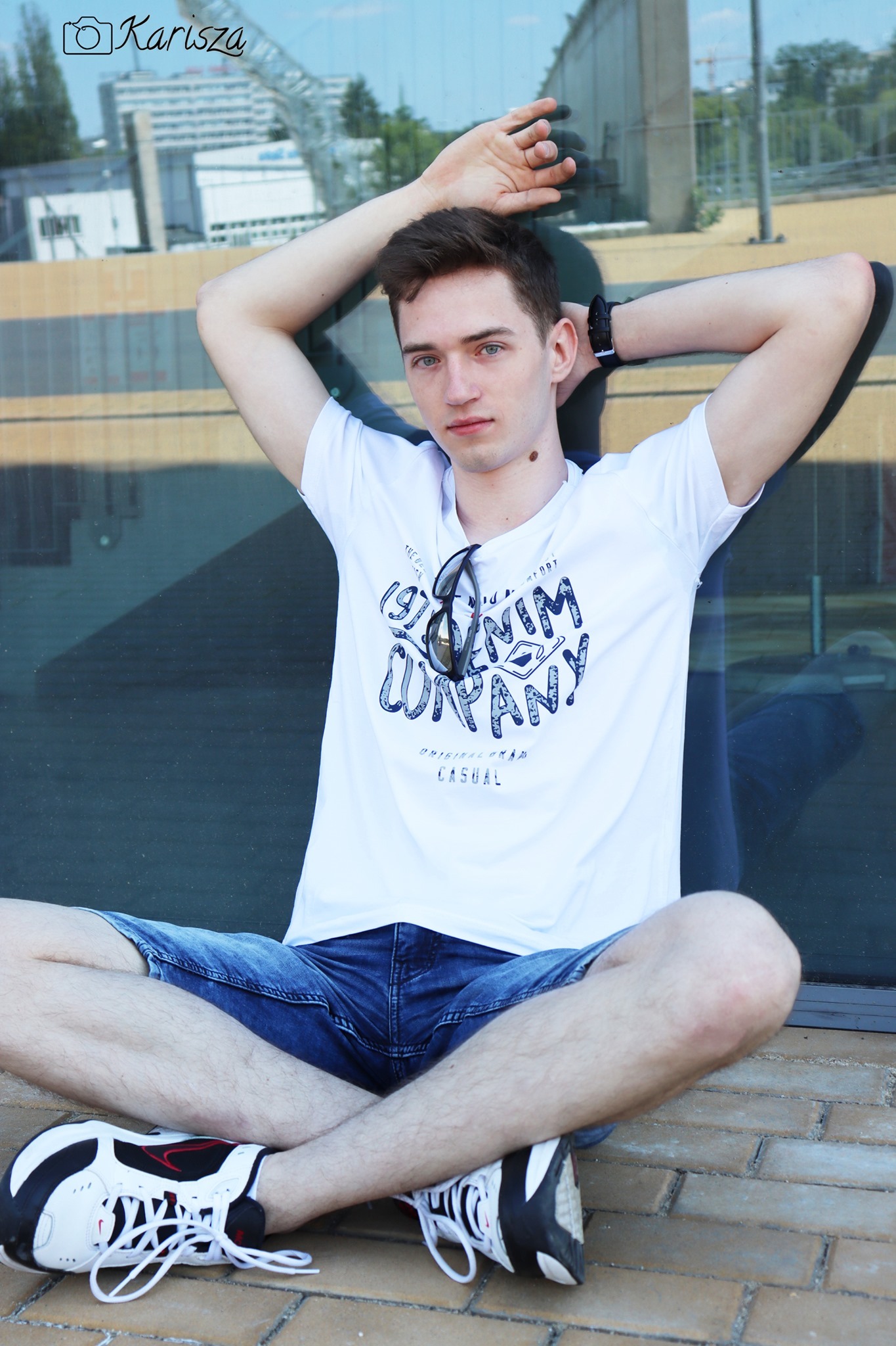 Młody mężczyzna w białej koszulce i jeansowych szortach siedzi skrzyżnie na ceglanej powierzchni, opierając się o szklaną ścianę, z okularami przeciwsłonecznymi zawieszonymi na koszulce i zegarkiem...