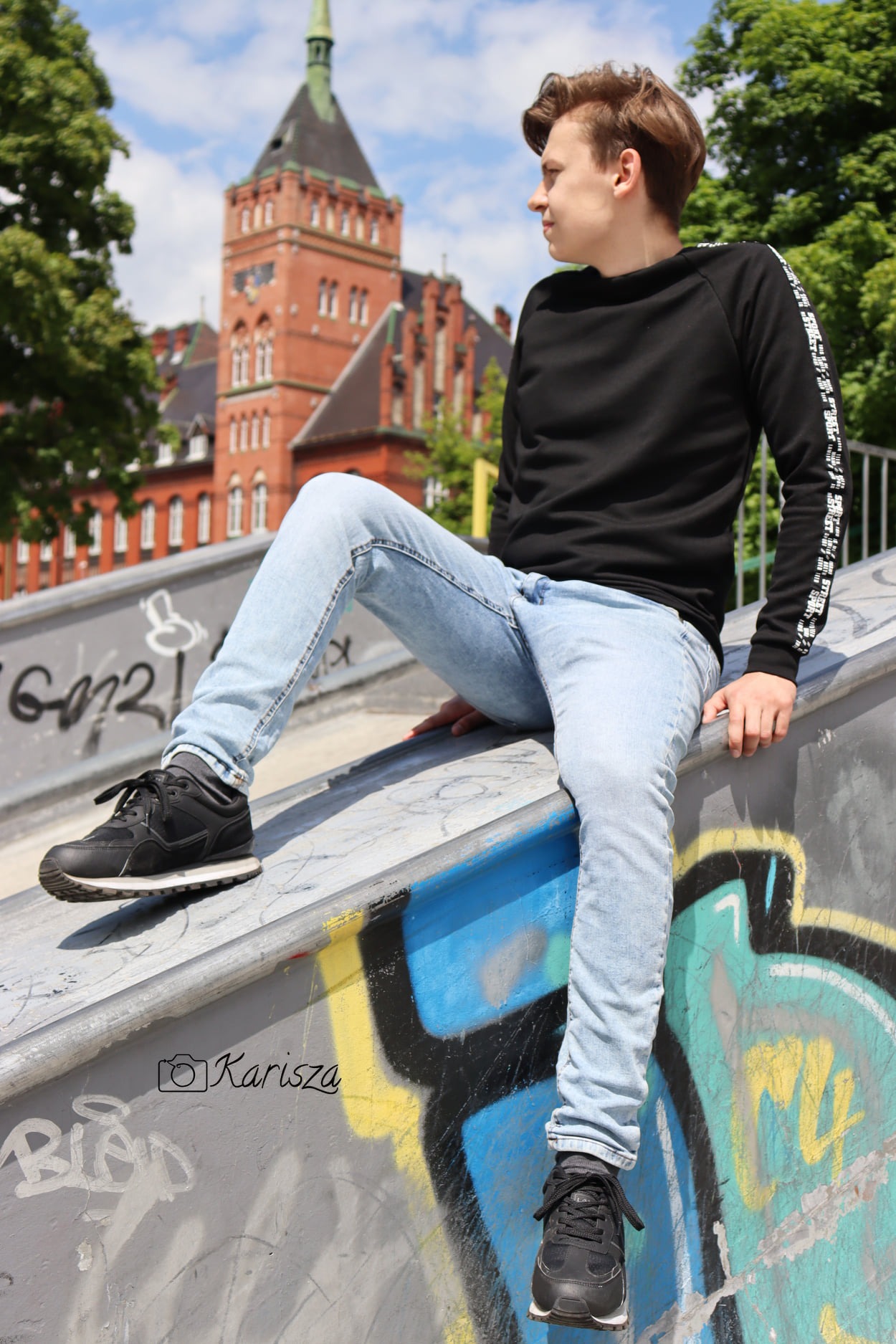 Młody mężczyzna w czarnej bluzie i jasnych jeansach siedzi na krawędzi skateparku z graffiti, w tle ceglany budynek z wieżą.