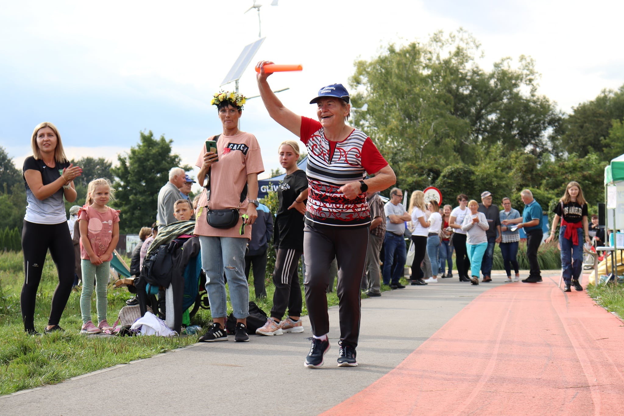 Uśmiechnięta kobieta w sportowym stroju z uniesioną ręką trzymająca pomarańczowy przedmiot, prawdopodobnie podczas biegu lub zawodów, na tle kibiców i zieleni.