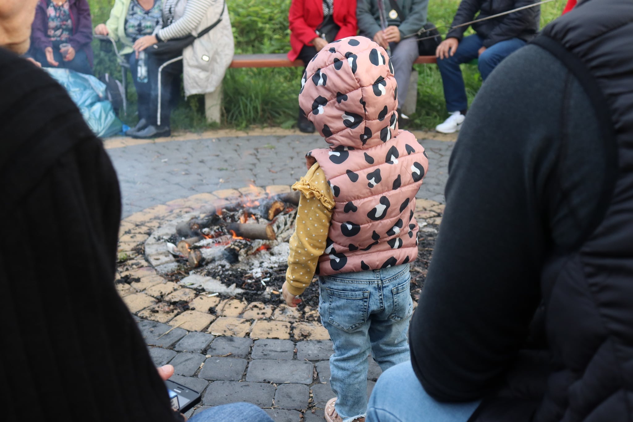 Mała dziewczynka w różowej kamizelce z kapturem w czarne cętki stoi tyłem przy ognisku otoczonym kamieniami, w tle siedzą osoby na ławce.