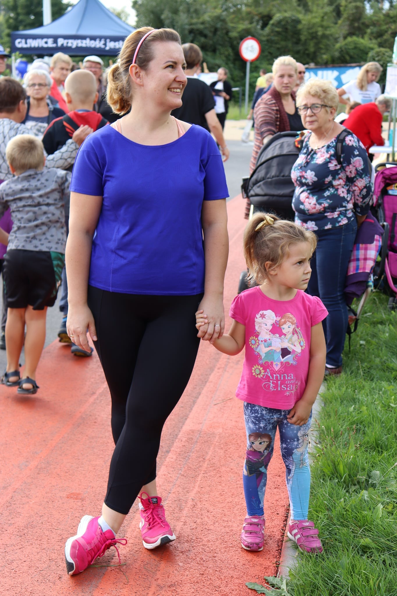 Kobieta w niebieskiej koszulce i czarnych legginsach trzyma za rękę małą dziewczynkę w różowej koszulce z postaciami z bajki Frozen, oboje stoją na czerwonym chodniku.