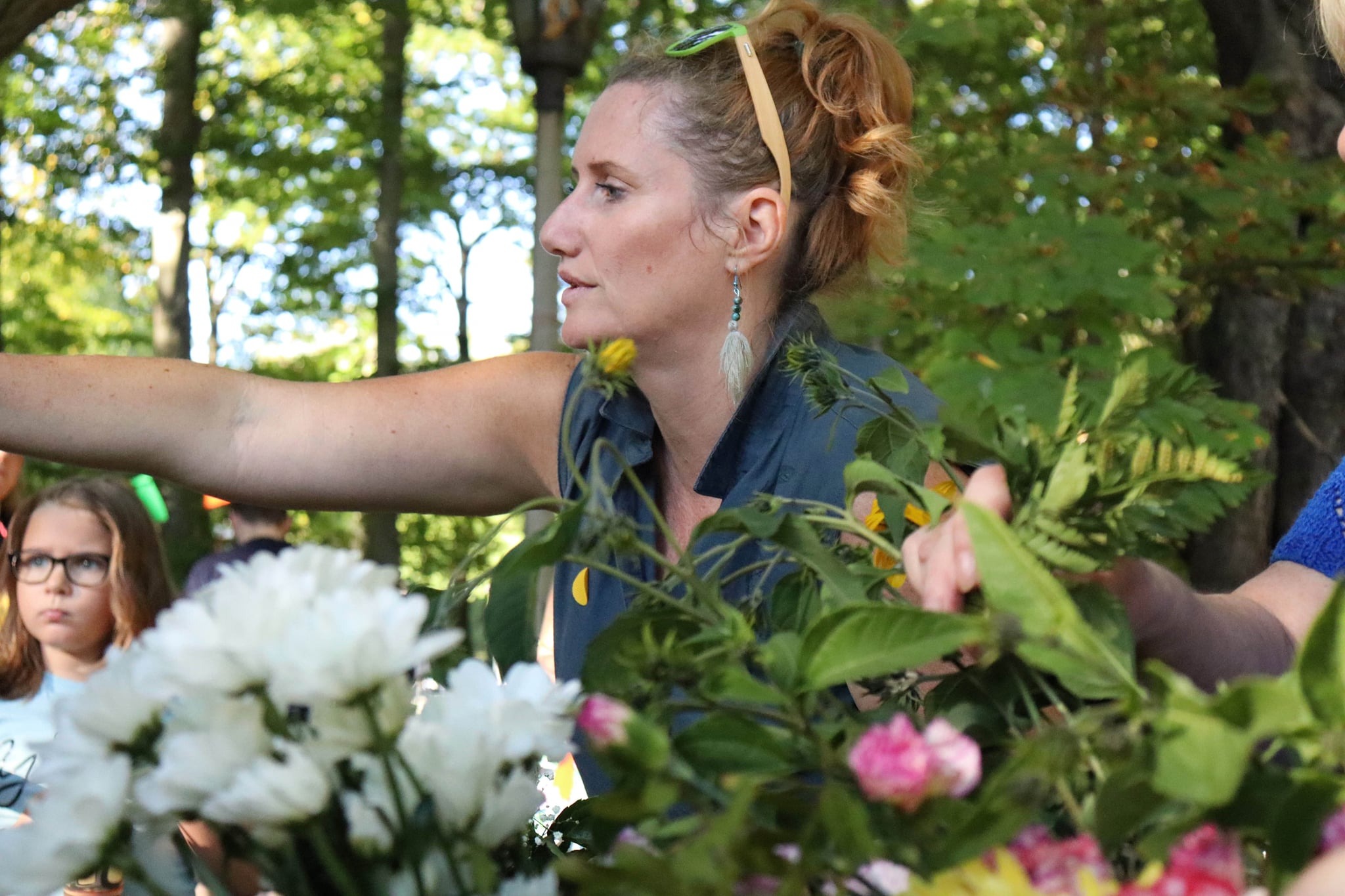 Kobieta z włosami upiętymi spinką w okularach na głowie, w trakcie tworzenia kompozycji kwiatowej na tle parku, z widoczną dziewczynką w okularach po lewej stronie.