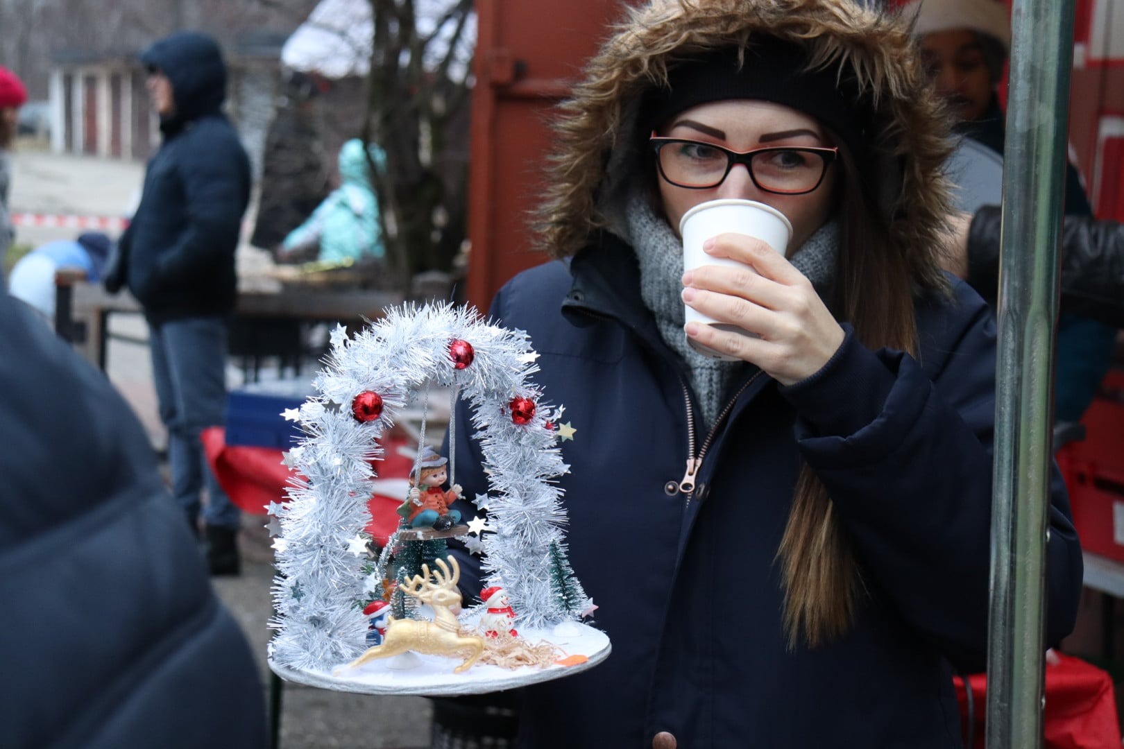 Kobieta w okularach i kurtce z kapturem pije z papierowego kubka, trzymając świąteczną dekorację z figurką dziecka na huśtawce, renifera i choinki.