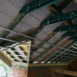 Konstrukcja podwieszanego sufitu z profili metalowych, widoczne drewniane elementy więźby dachowej, ocieplenie i membrana dachowa, surowy mur z cegły dziurawki w tle.
