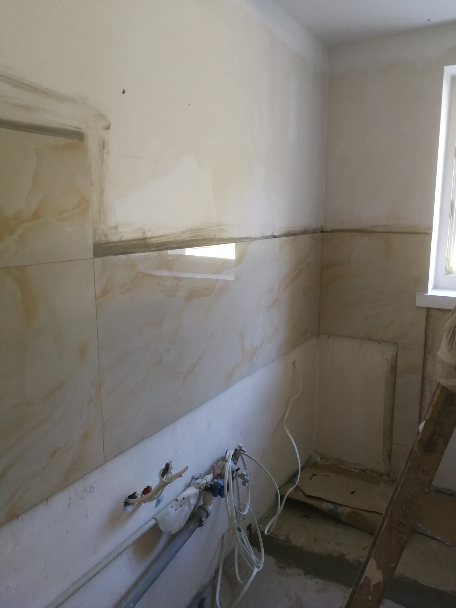 Fragment kuchni w trakcie remontu: częściowo położone marmurkowe płytki na ścianie, odsłonięte przewody i rury, widoczne ślady po pracach budowlanych, drabina oparta o ścianę.