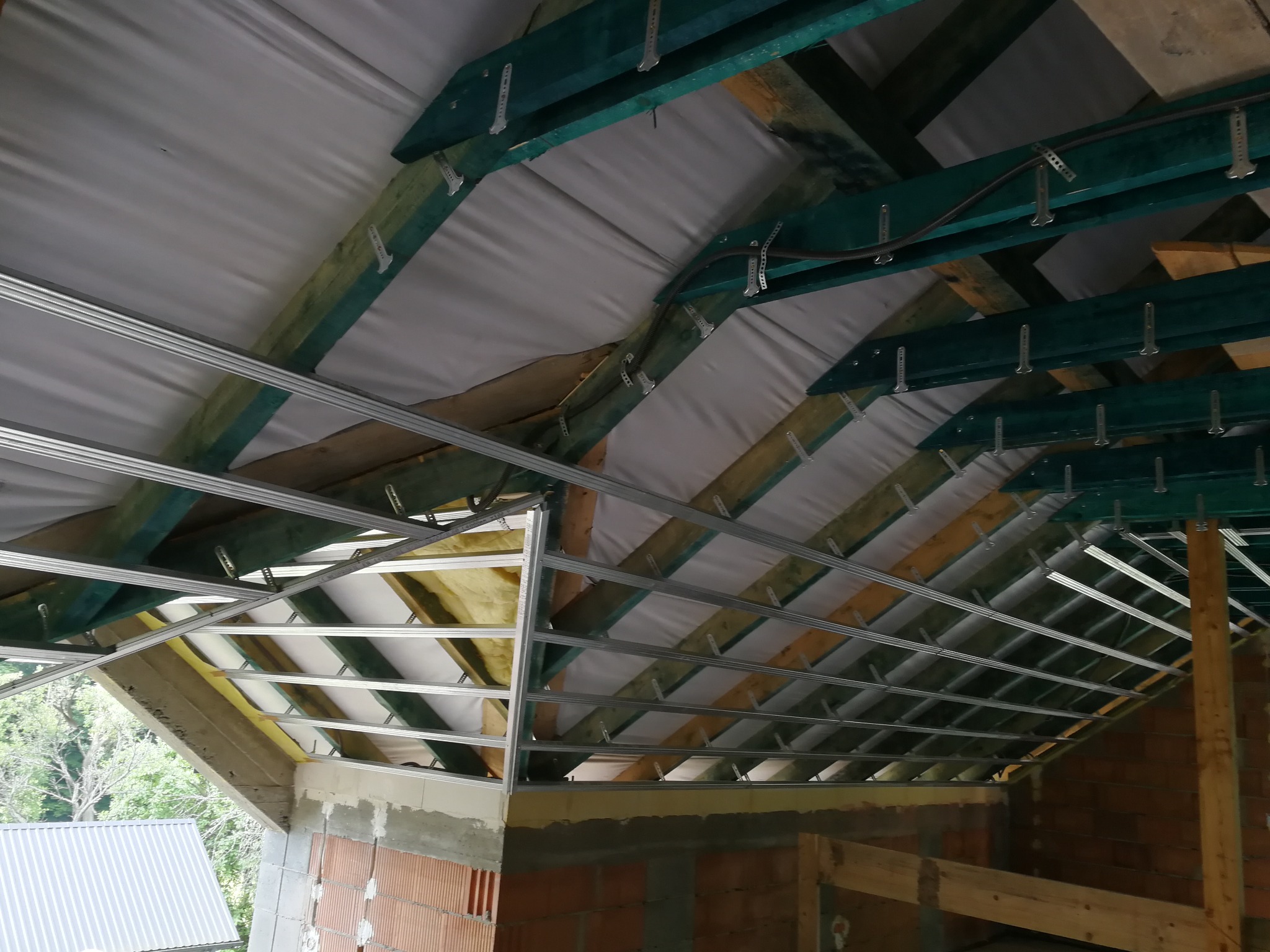 Konstrukcja podwieszanego sufitu z profili metalowych, widoczne drewniane elementy więźby dachowej, ocieplenie i membrana dachowa, surowy mur z cegły dziurawki w tle.