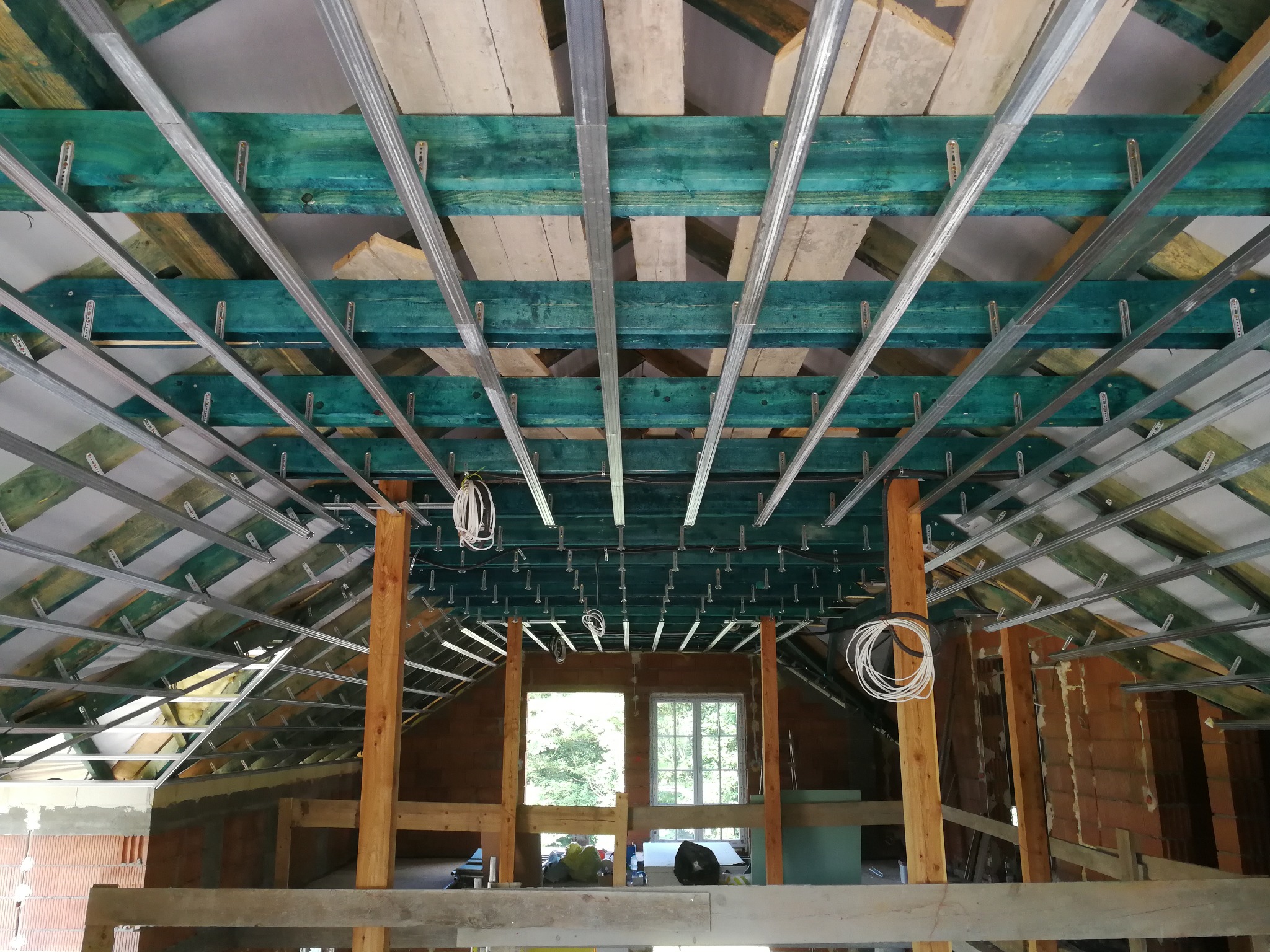 Metalowa konstrukcja podwieszanego sufitu zamontowana na drewnianych belkach w surowym wnętrzu poddasza, widoczne okna i instalacja elektryczna.