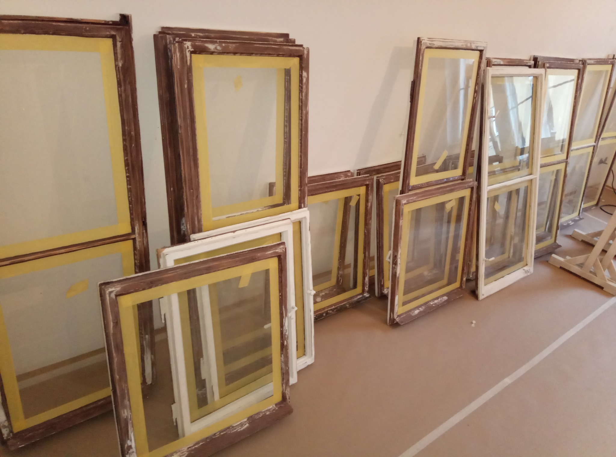 Zdjęcie przedstawia przygotowane do odnowienia drewniane ramy okienne, zabezpieczone taśmą malarską, ułożone na podłodze i oparte o ścianę.