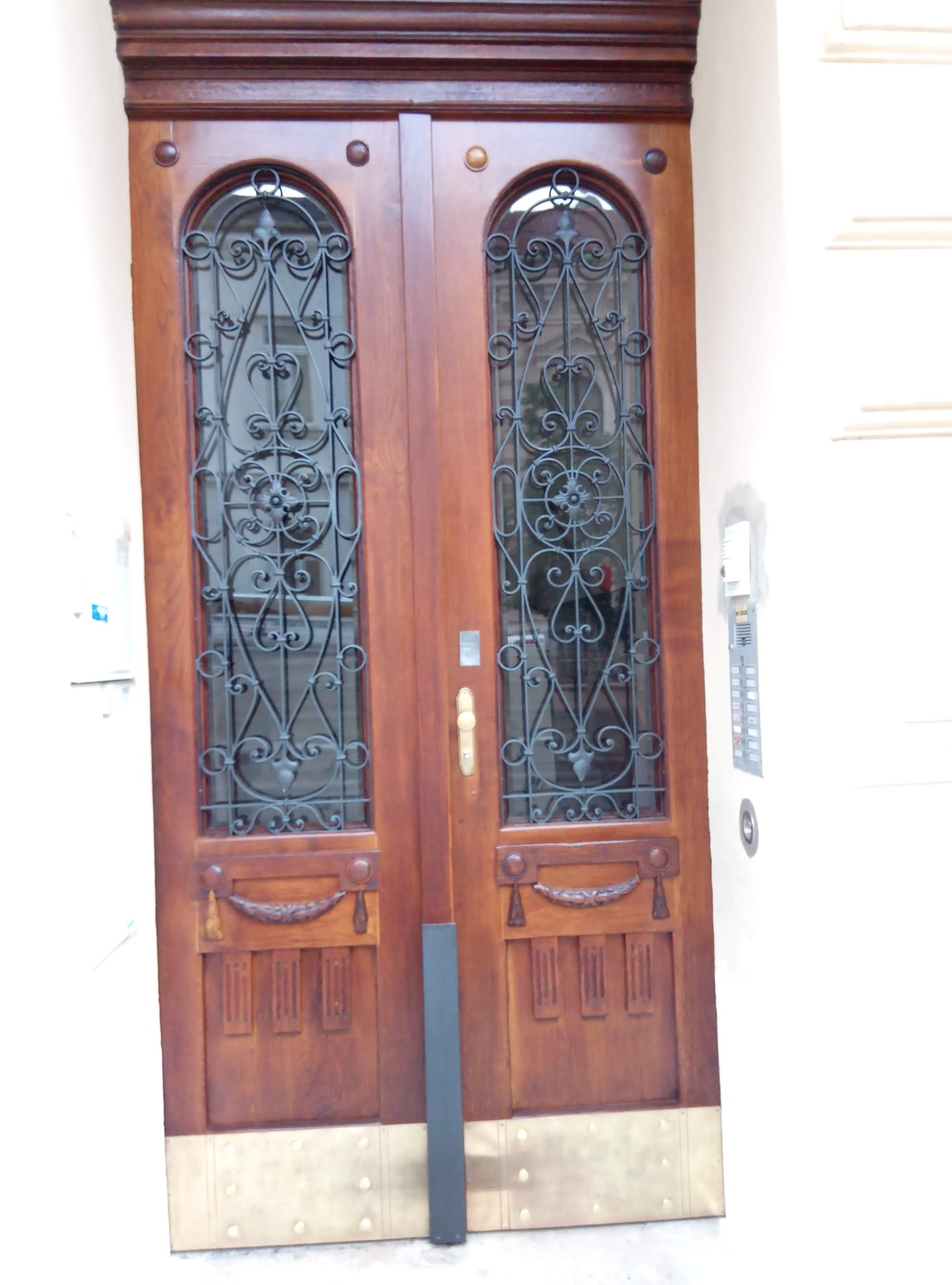 Odnowione, dwuskrzydłowe drzwi wejściowe z ciemnego drewna, zdobione metalową kratą o dekoracyjnych wzorach i mosiężnymi elementami u dołu.