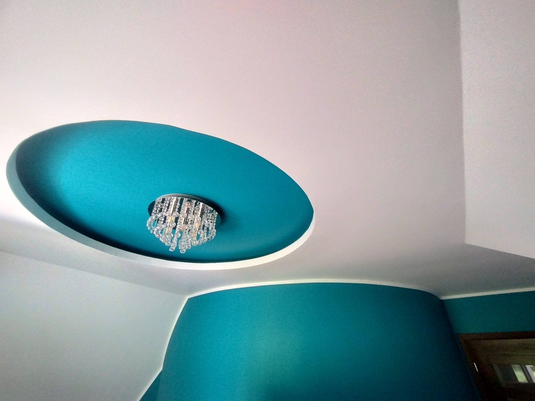 Turkusowe akcenty w nowoczesnym wnętrzu w Nowym Wiśniczu: okrągła wnęka w suficie z kryształowym żyrandolem i fragment ściany w tym samym kolorze.