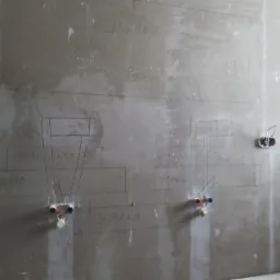 Surowa ściana łazienki w trakcie przygotowań do położenia płytek, z zaznaczonymi miejscami podłączeń wodnych i elektrycznych oraz odręcznymi notatkami na ścianie.