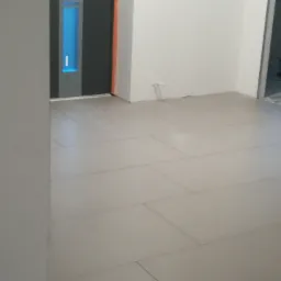 Ułożone, jasne płytki podłogowe w korytarzu, widoczna instalacja elektryczna przy ścianie, fragment drzwi z oświetleniem LED oraz sprzęt budowlany w tle przez otwarte drzwi.