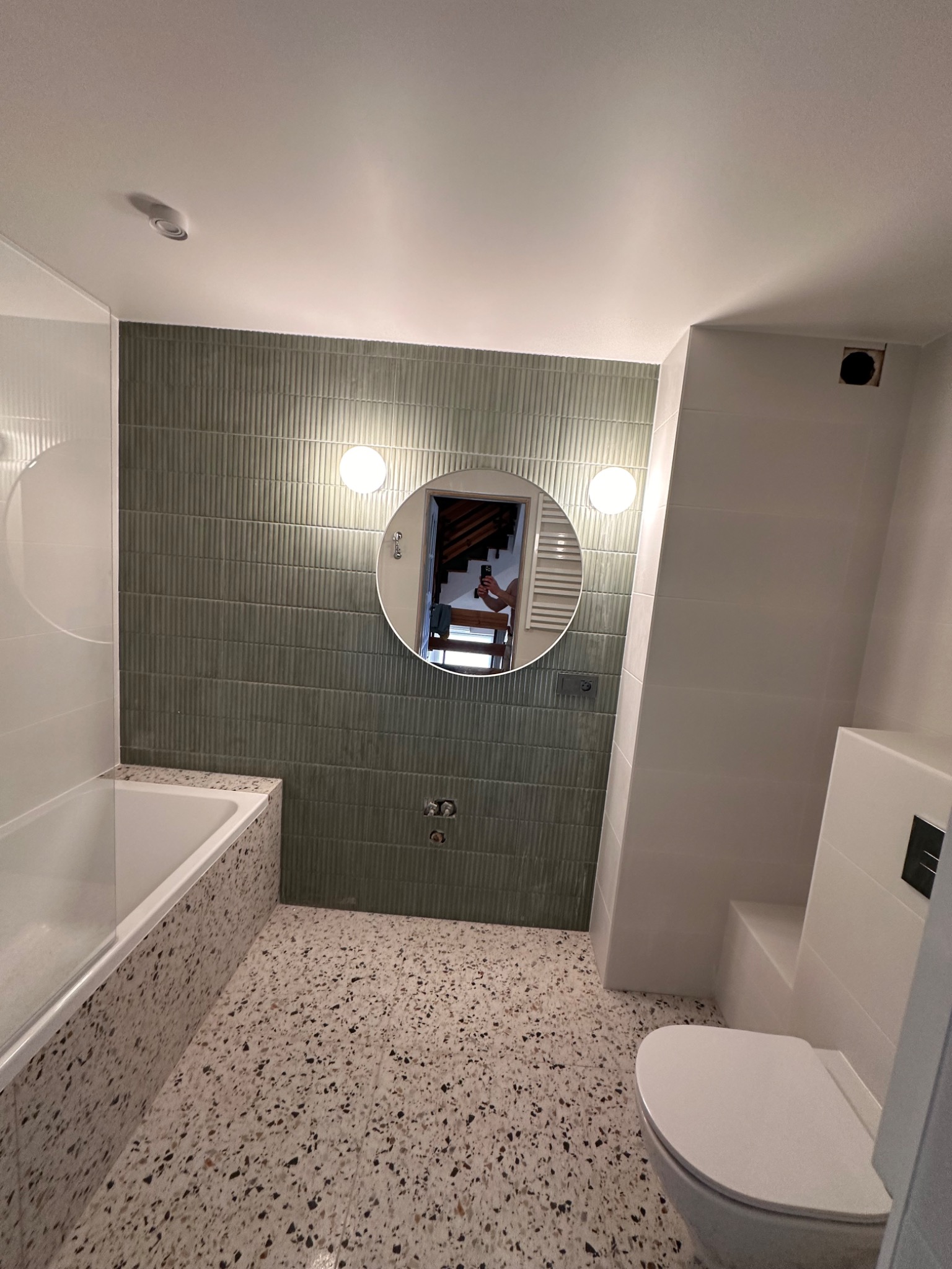 Nowoczesna łazienka z wanną obudowaną mozaiką lastryko, zielone płytki na ścianie za okrągłym lustrem z oświetleniem, toaleta podwieszana, podłoga lastryko.