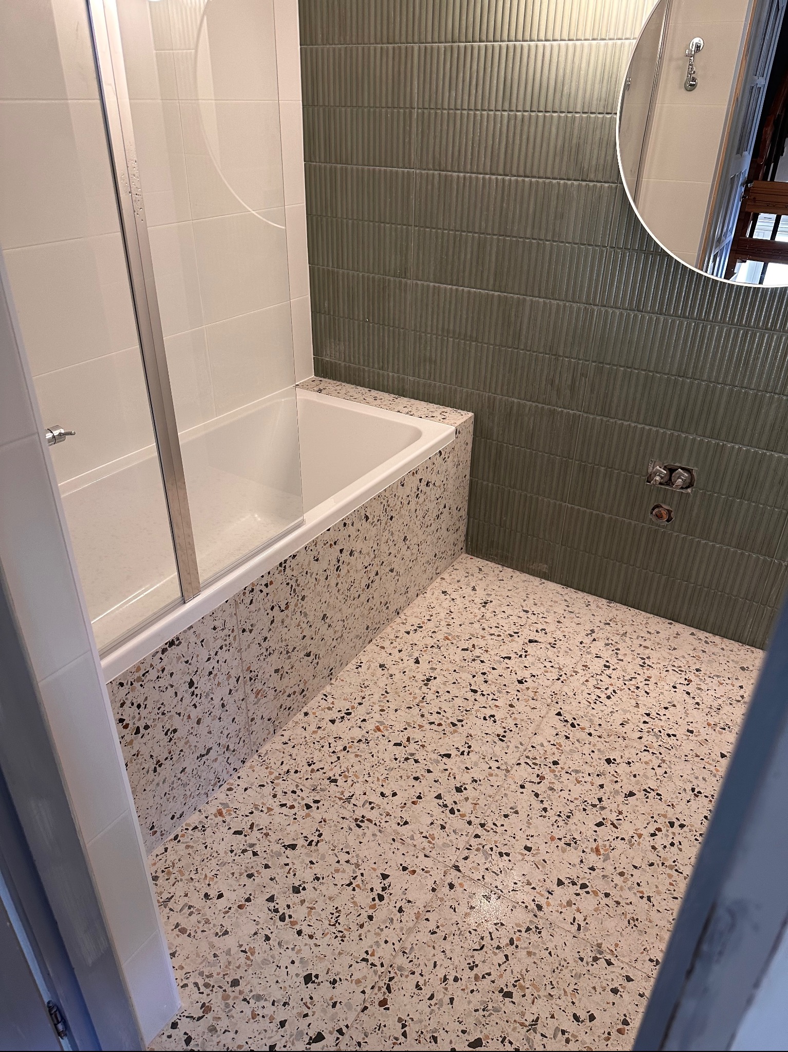 Nowoczesna łazienka z wanną za szklaną ścianką, wykończona płytkami imitującymi lastryko na podłodze i obudowie wanny, oraz pionowymi, żłobionymi płytkami w odcieniach zieleni na ścianie. Widoczne...