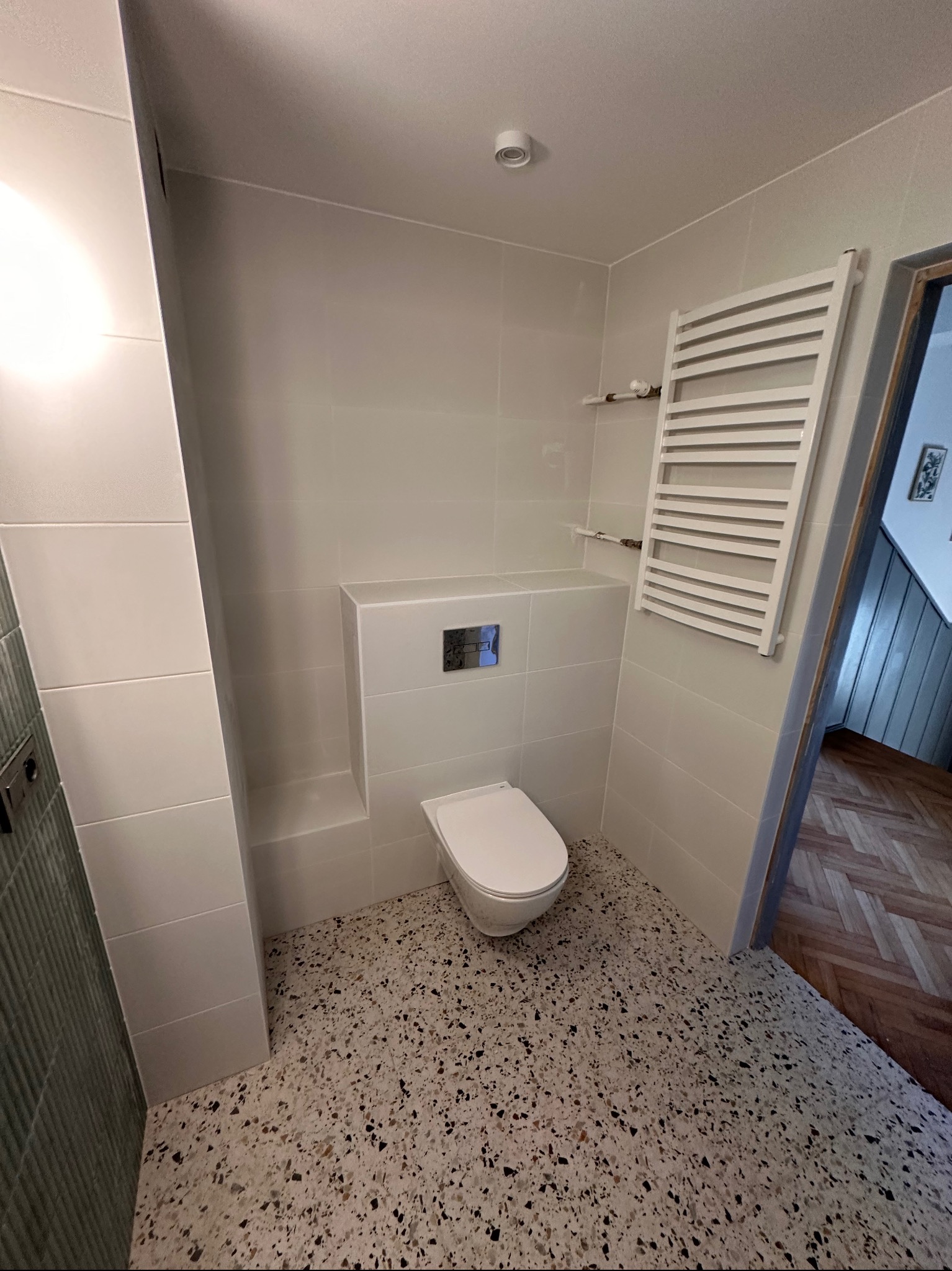 Nowoczesna łazienka po remoncie: białe płytki ścienne, podłoga lastryko, wisząca toaleta z chromowanym przyciskiem, grzejnik drabinkowy i fragment parkietu w przedpokoju