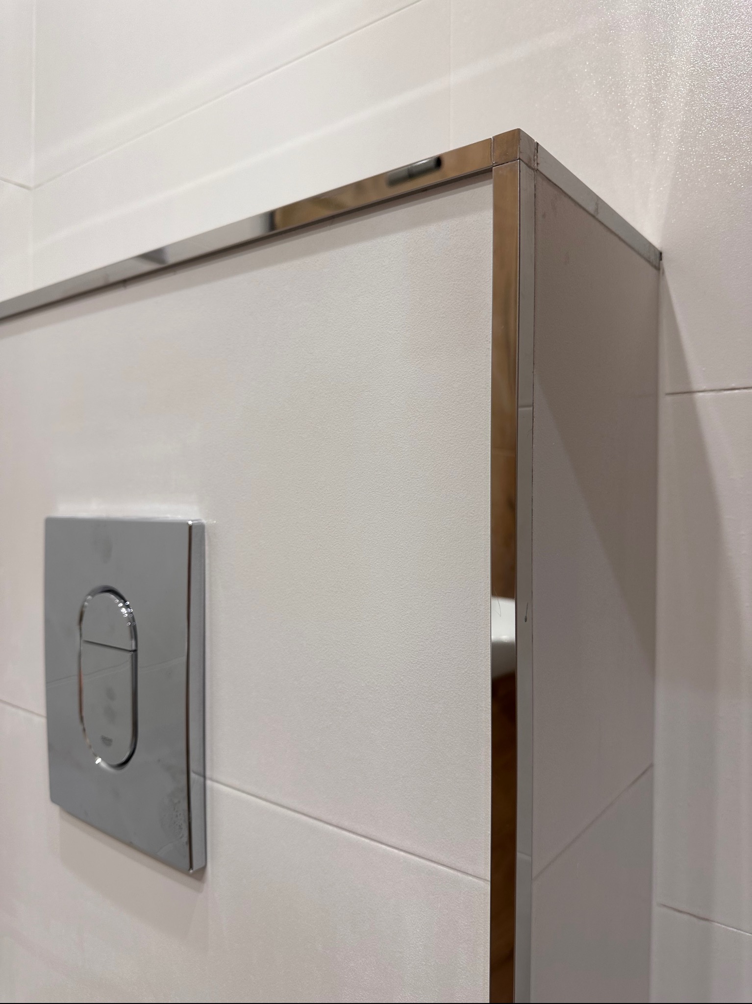 Biała ściana z płytek z chromowanym przyciskiem spłuczki i narożnym wykończeniem, widoczny fragment toalety w odbiciu.