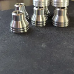 Sześć precyzyjnie obrobionych, aluminiowych elementów o kształcie stożka z rowkami, ułożonych na ciemnej powierzchni. W tle polerka z okrągłą szczotką i żółty akumulatorowy wkrętak.