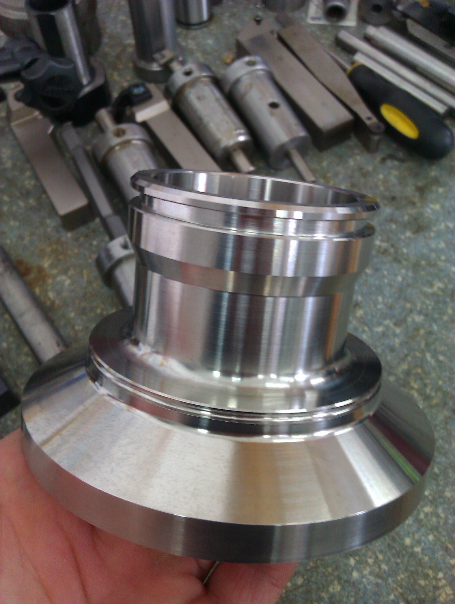 Precyzyjnie obrobiony, błyszczący element metalowy o złożonym kształcie, trzymany w dłoni, z widocznymi narzędziami i innymi metalowymi częściami w tle.