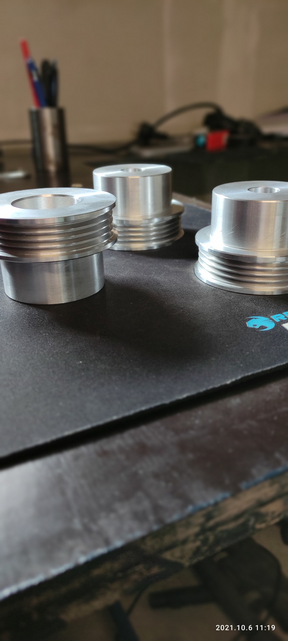 Trzy precyzyjnie obrobione, srebrne elementy z aluminium z rowkami, leżące na czarnej podkładce na biurku, w tle widoczny pojemnik z długopisami i kablami.