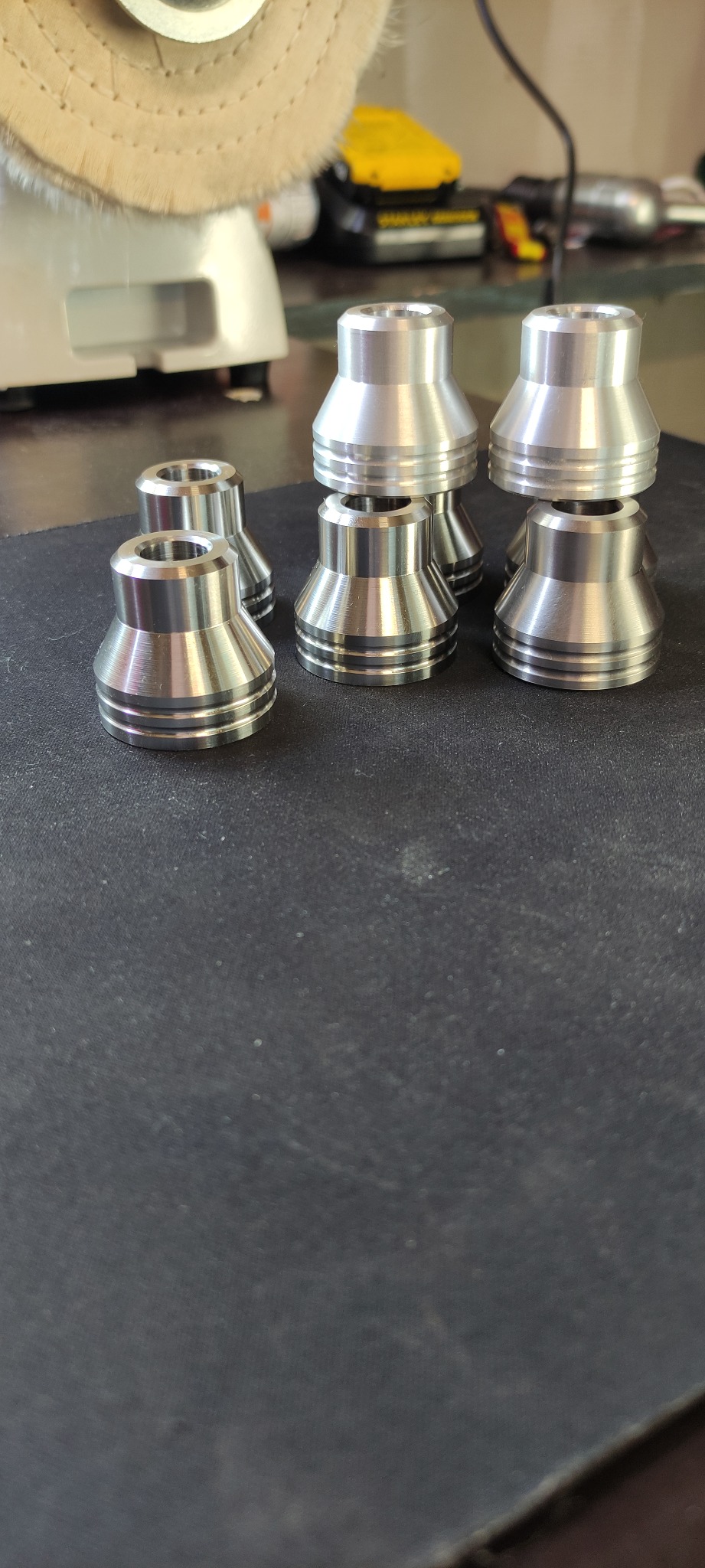 Sześć precyzyjnie obrobionych, aluminiowych elementów o kształcie stożka z rowkami, ułożonych na ciemnej powierzchni. W tle polerka z okrągłą szczotką i żółty akumulatorowy wkrętak.