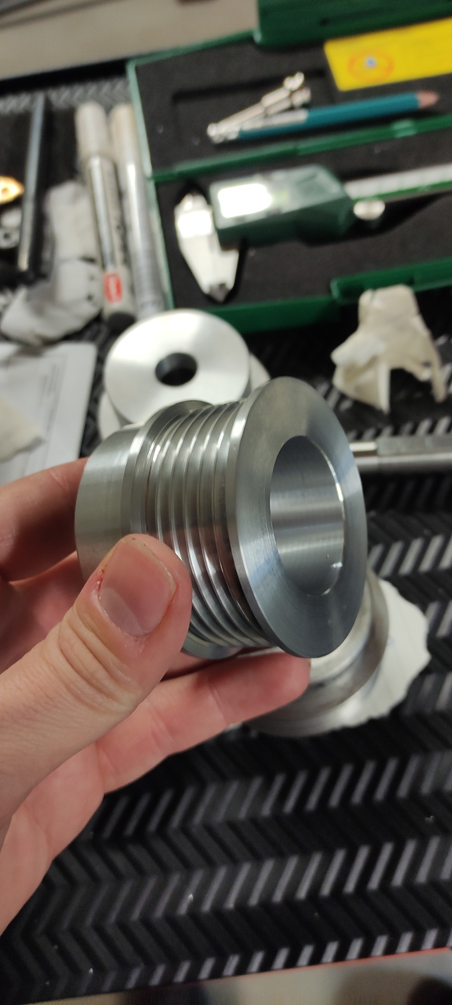 Precyzyjnie wykonany, metalowy element z rowkami, trzymany w dłoni, w tle narzędzia pomiarowe i inne metalowe detale.