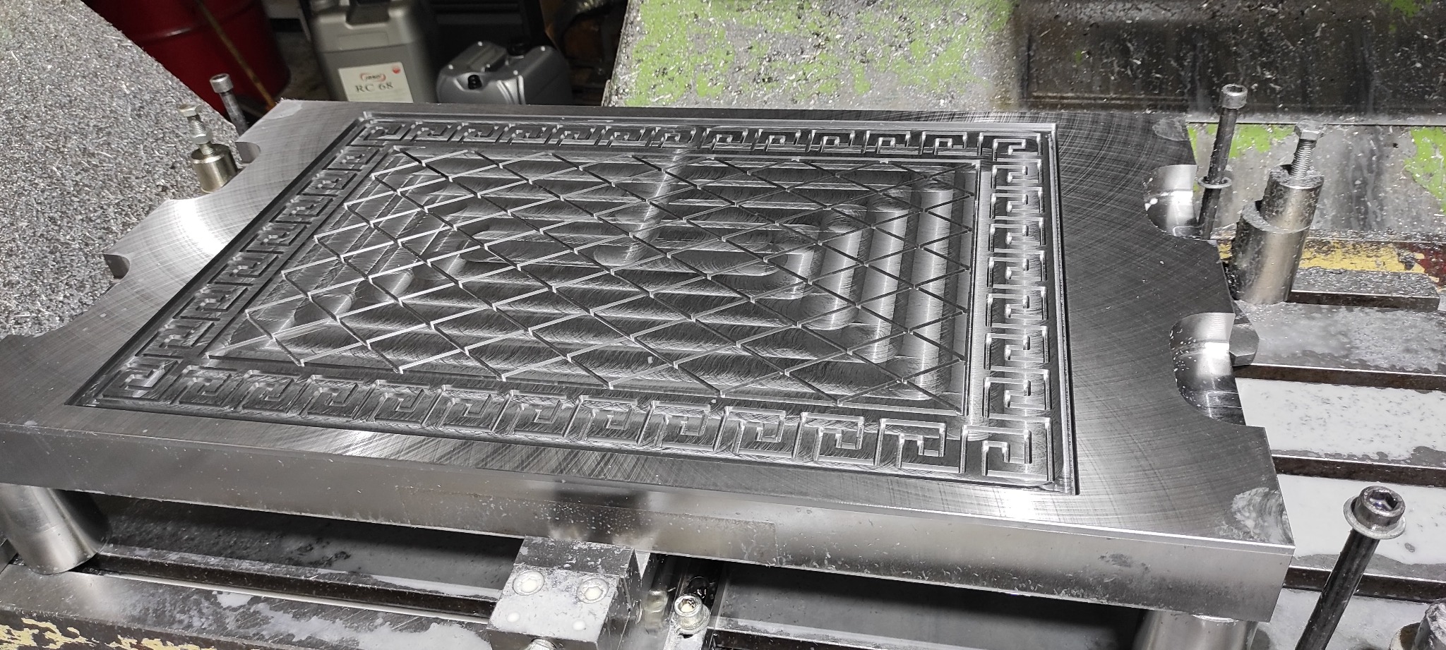 Metalowa płyta z wyfrezowanym geometrycznym wzorem, zamocowana na obrabiarce CNC, widoczne ślady obróbki i wióry.