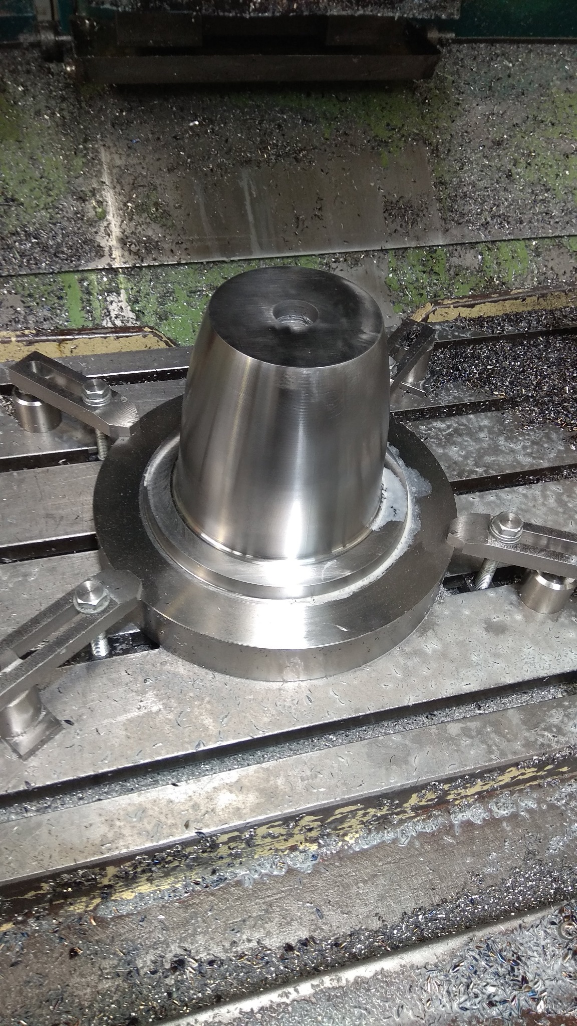 Metalowy element w trakcie obróbki na maszynie CNC, zabezpieczony uchwytami, widoczne wióry i płyn chłodzący.