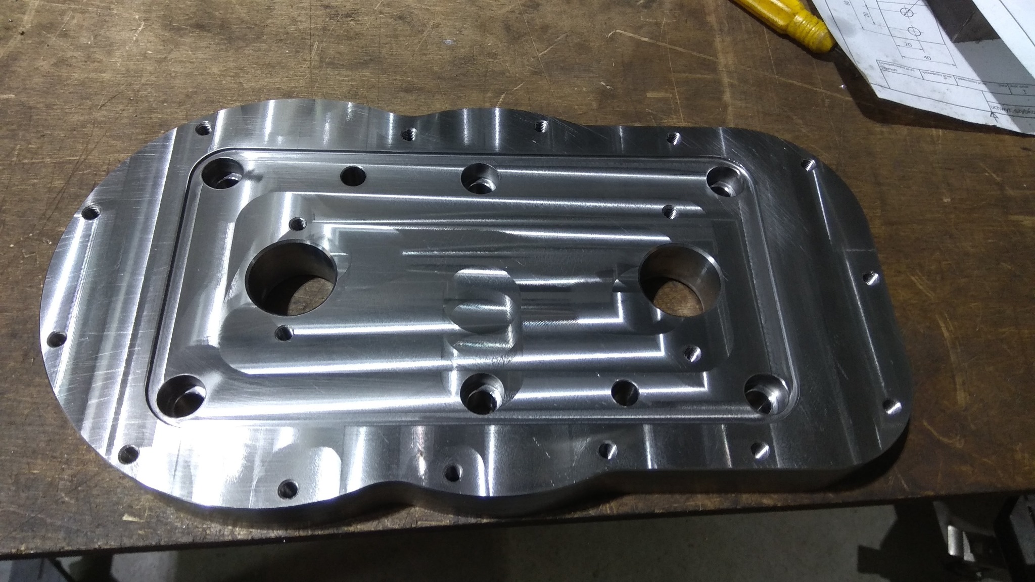 Błyszczący, precyzyjnie obrobiony element metalowy z licznymi otworami i frezowaniami, leżący na drewnianym stole warsztatowym, obok widoczny fragment rysunku technicznego i żółty śrubokręt.