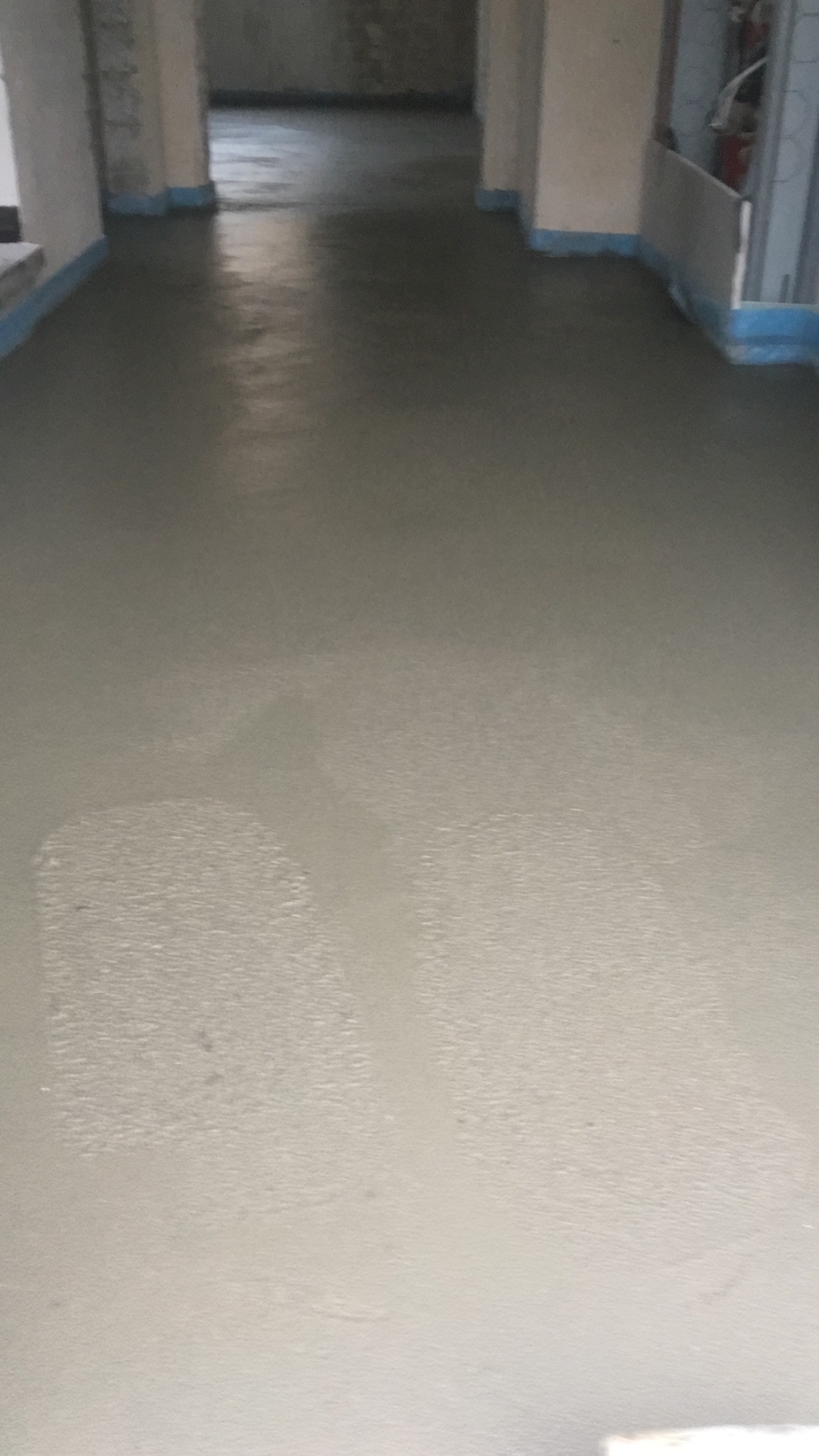 Świeżo wylana, gładka posadzka jastrychowa w surowym wnętrzu budynku, zabezpieczona niebieską folią przy ścianach.