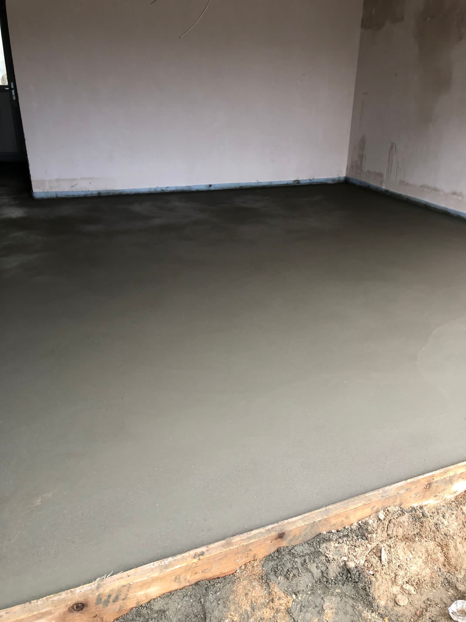 Świeżo wylana, gładka posadzka jastrychowa w surowym pomieszczeniu z widocznymi deskami szalunkowymi przy krawędzi.