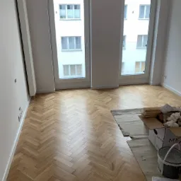 Parkiet ułożony we wzór jodełki w jasnym pokoju z dwoma oknami, widoczne materiały budowlane i puszka farby w rogu, ściany pomalowane na biało.