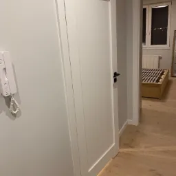 Białe drzwi wewnętrzne z czarną klamką, widoczne z perspektywy korytarza, z fragmentem parkietu i wnętrzem sypialni w tle, z interkomem na ścianie.