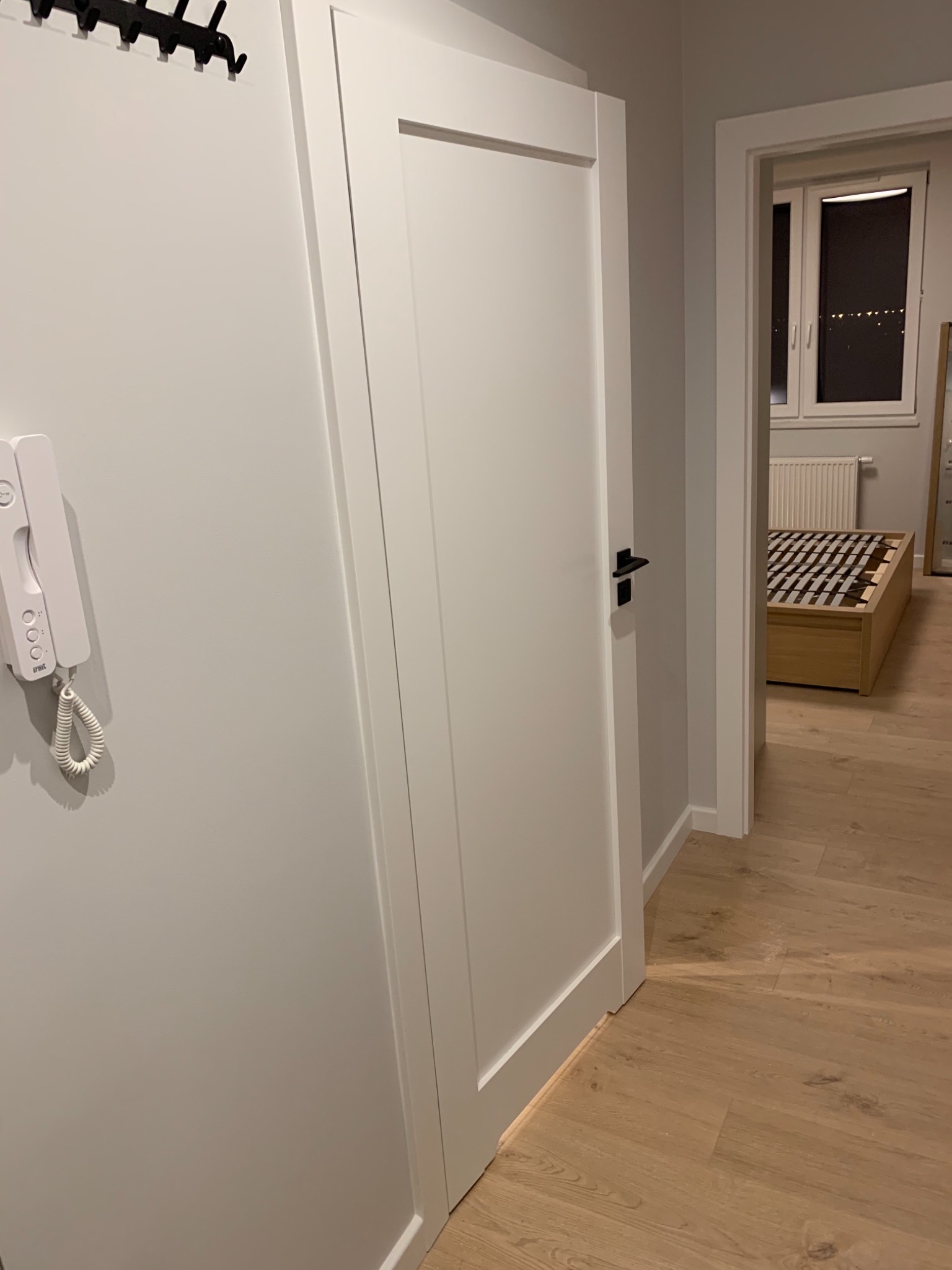 Białe drzwi wewnętrzne z czarną klamką, widoczne z perspektywy korytarza, z fragmentem parkietu i wnętrzem sypialni w tle, z interkomem na ścianie.