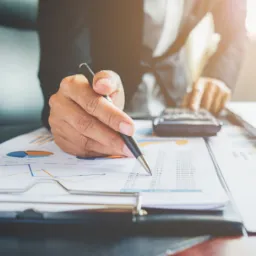 Osoba w garniturze analizuje dokumenty z wykresami i tabelami, używając kalkulatora i długopisu.