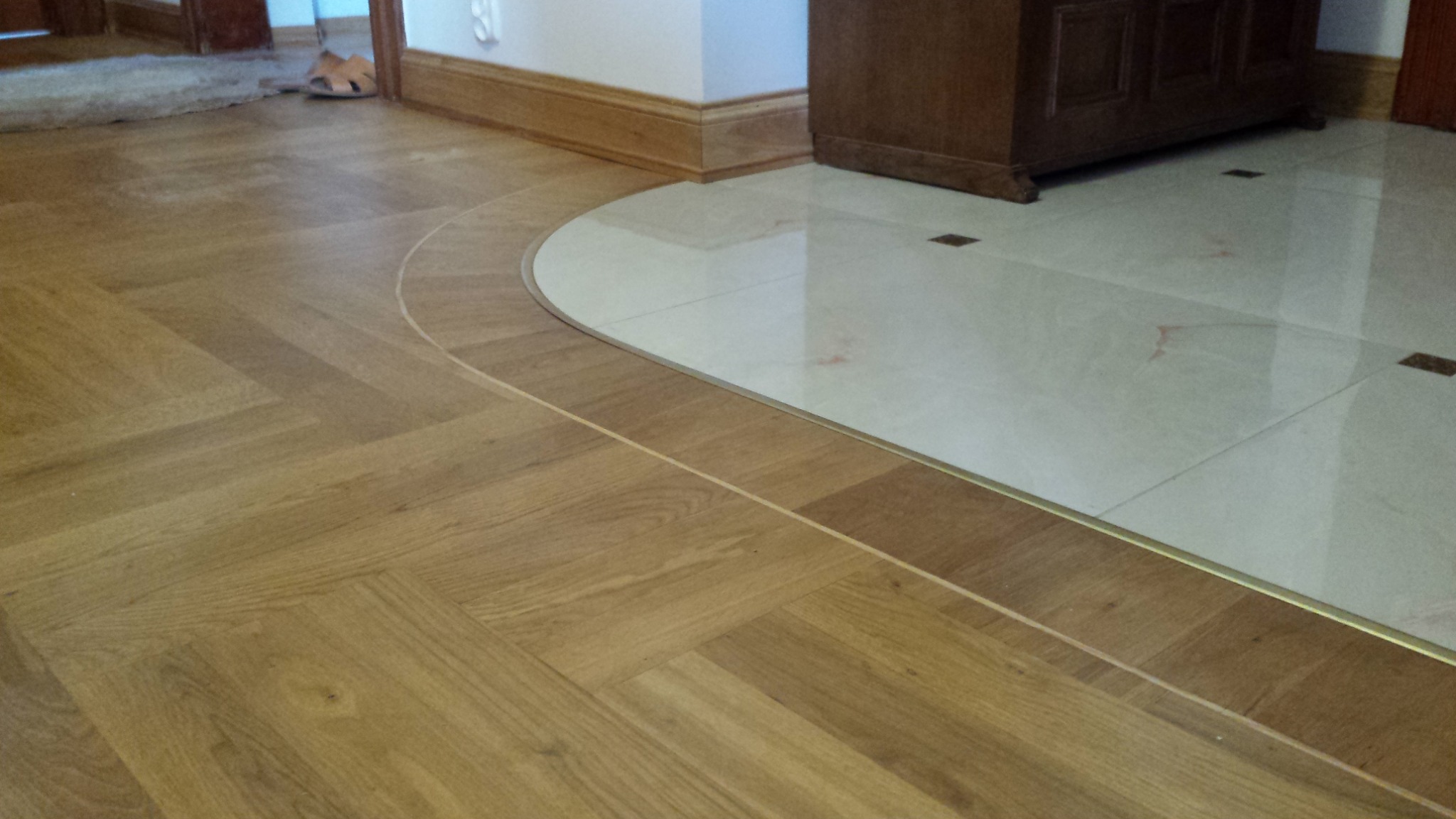 Połączenie parkietu ułożonego w jodełkę z jasnymi płytkami ceramicznymi przy użyciu listwy dylatacyjnej w kolorze złotym, widoczne fragmenty mebli i drzwi.