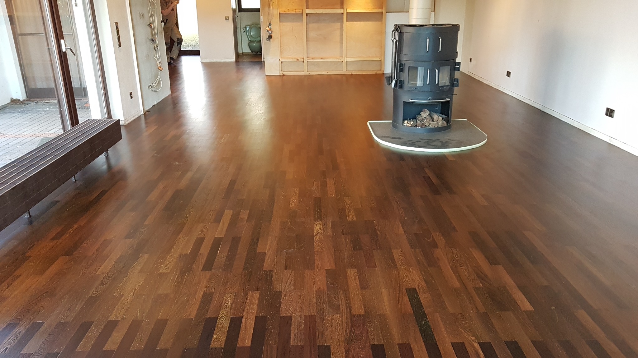 Ciemnobrązowa, lakierowana podłoga parkietowa ułożona w jodełkę francuską, z widocznym czarnym piecem wolnostojącym i minimalistyczną ławką w tle remontowanego pomieszczenia.