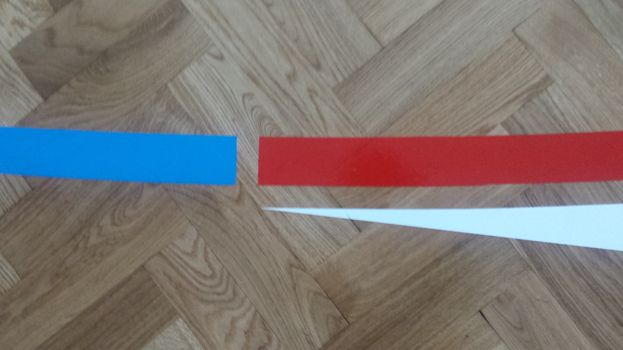 Parkiet ułożony w jodełkę z trzema kolorowymi pasami taśmy malarskiej: niebieskim, czerwonym i białym, tworzącymi kompozycję abstrakcyjną.