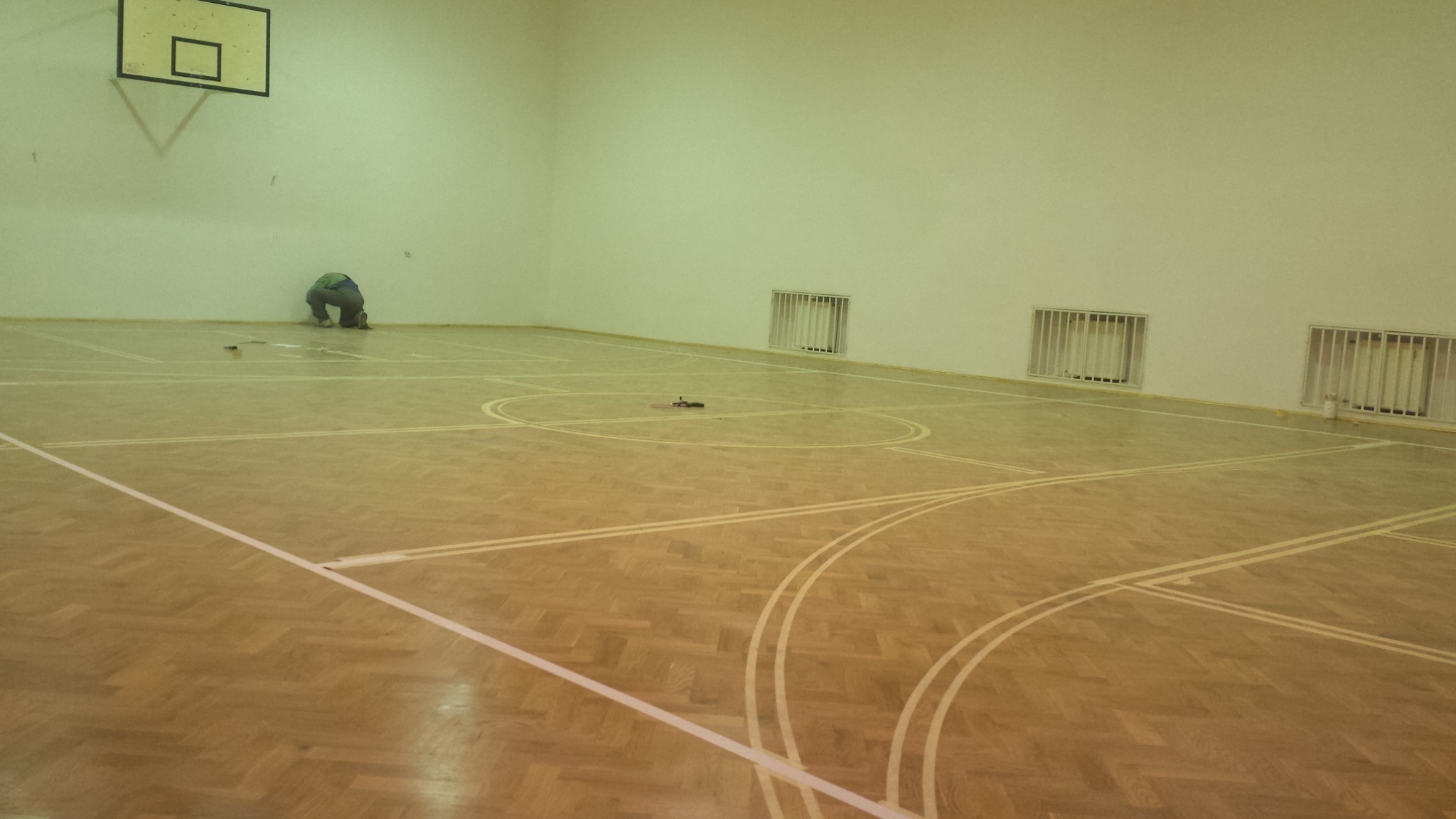 Sala gimnastyczna z parkietem w jodełkę, na którym widoczne są linie boiska; osoba w kucki przy ścianie, prawdopodobnie wykonująca prace wykończeniowe, oraz narzędzia na podłodze.