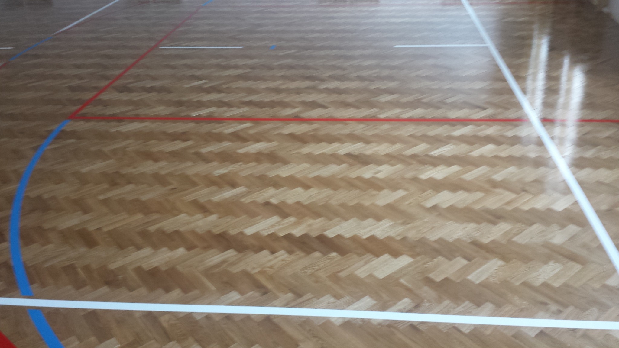 Parkiet ułożony w jodełkę na podłodze hali sportowej, z widocznymi liniami boiska w kolorach białym, czerwonym i niebieskim.