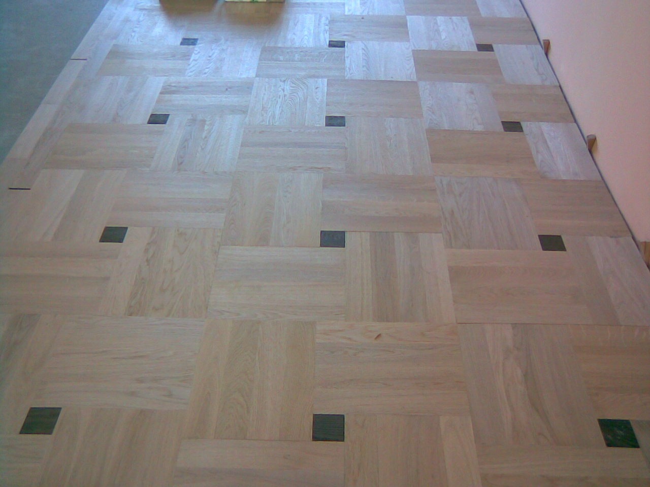 Ułożony parkiet z jasnego drewna w kwadraty z ciemnymi, kwadratowymi wstawkami, widoczne szczeliny między niektórymi elementami.