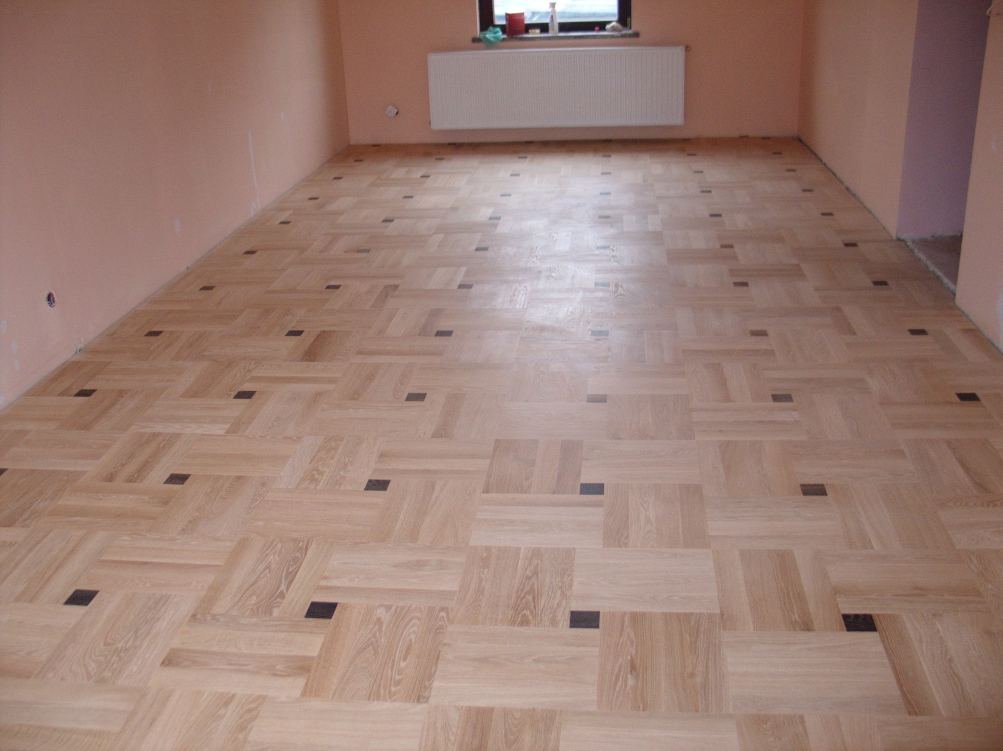 Jasny parkiet ułożony we wzór szachownicy z ciemnymi akcentami w przestronnym, pomalowanym na różowo pokoju z białym grzejnikiem pod oknem.