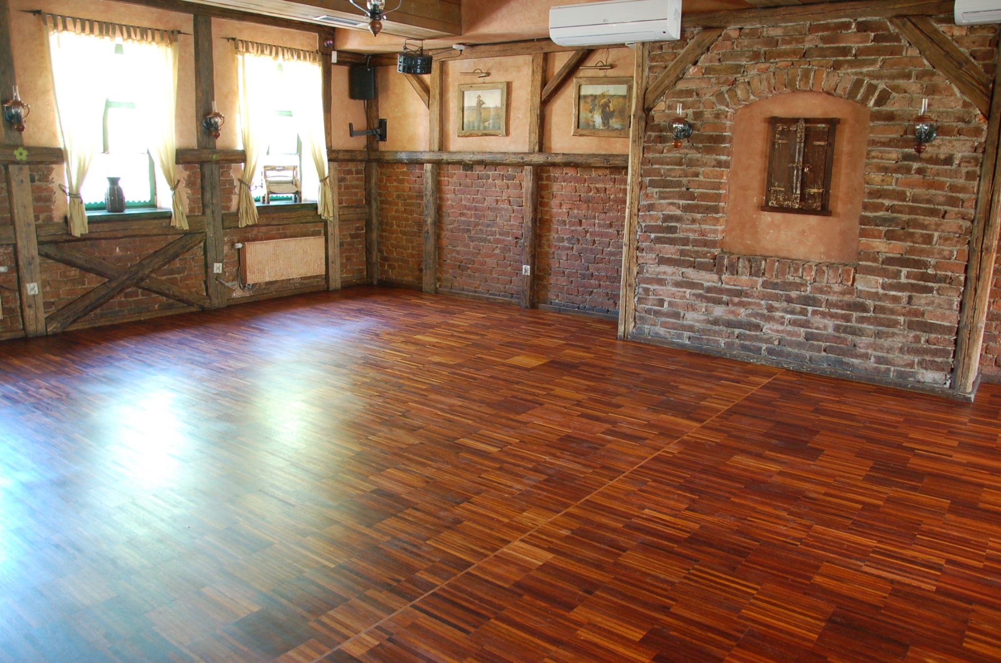 Wnętrze sali z parkietem ułożonym w jodełkę, ceglane ściany z drewnianymi belkami, okna z zasłonami i dekoracjami.