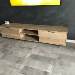 Drewniana szafka RTV w odcieniach dębu, zawieszona na szarej ścianie z efektem betonu, z widocznym fragmentem telewizora u góry i szarą podłogą imitującą kamień.