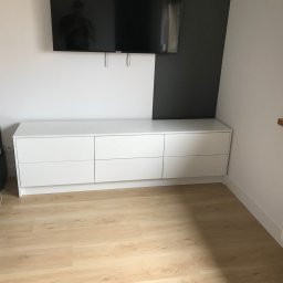 Dawid Brociek - Biała komoda z sześcioma szufladami w sypialni, ustawiona pod telewizorem na ścianie pomalowanej w dwóch kolorach, obok łóżko z szarym zagłówkiem.