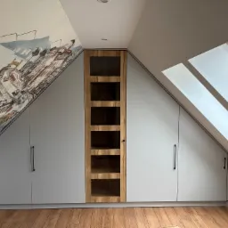 Szafa wnękowa na poddaszu ze ścianą ozdobioną malowidłem. Fronty w kolorze szarym, środkowa część z drewnianą ramą i przeszkleniami. Widoczne okno dachowe.